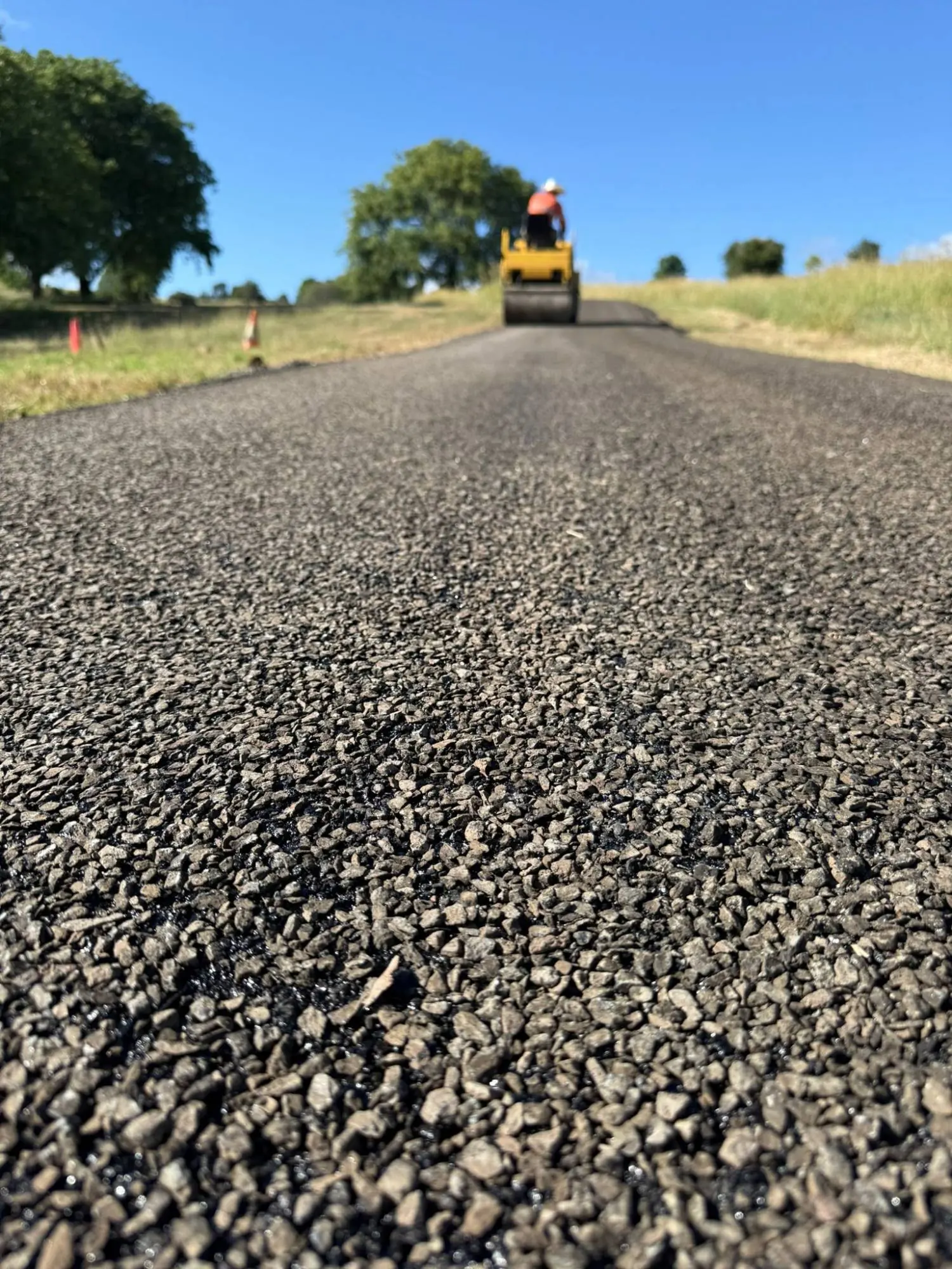 A freshly spray-sealed asphalt road with loose dark aggregate in the foreground, a road roller working in the distance.